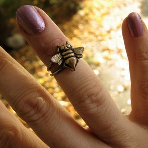 Può includere: Un anello d'ape d'argento con un designo dettagliato. L'anello è indossato su un dito con smalto per unghie viola.