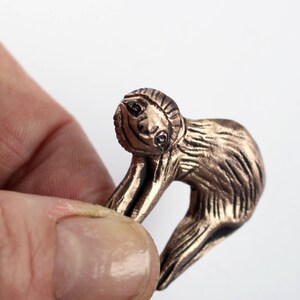 Pode incluir: Uma pequena figura de pregui&ccedil;a de cor bronze com uma superf&iacute;cie texturizada detalhada. A pregui&ccedil;a &eacute; representada em uma pose relaxada, pendurada com os bra&ccedil;os estendidos.