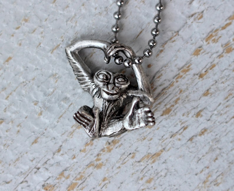 Puede incluir: Un collar de plata con un dije de mono. El mono est&aacute; sentado con los brazos levantados y las piernas colgando. El mono tiene una expresi&oacute;n juguetona en su rostro.