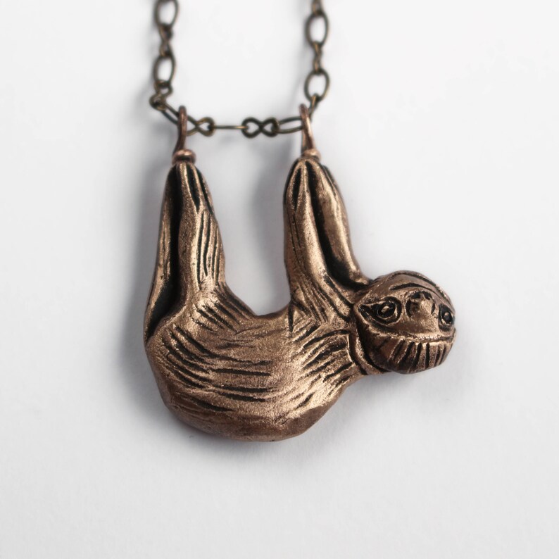 Pode incluir: Um colar com um pingente de pregui&ccedil;a de cor bronze. A pregui&ccedil;a est&aacute; pendurada de cabe&ccedil;a para baixo com os bra&ccedil;os e as pernas estendidos. O colar &eacute; feito de uma corrente marrom.