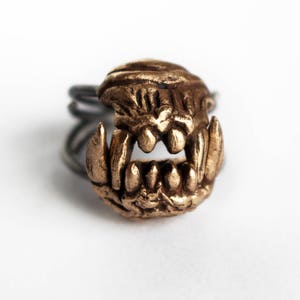 May include: A gold-toned ring with a detailed, toothy, monster-like design. The ring is made of metal and has a textured surface.