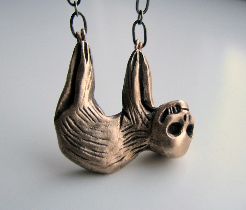 Pode incluir: Um pingente de bronze em forma de pregui&ccedil;a pendurado em uma corrente preta. A pregui&ccedil;a &eacute; esculpida em um estilo realista e detalhado, com os bra&ccedil;os e as pernas estendidos como se estivesse pendurado em um galho.