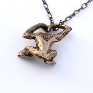 Puede incluir: Un collar con un colgante de bronce que presenta una figura estilizada de una persona en posici&oacute;n de cuclillas. El colgante est&aacute; unido a una cadena de plata.