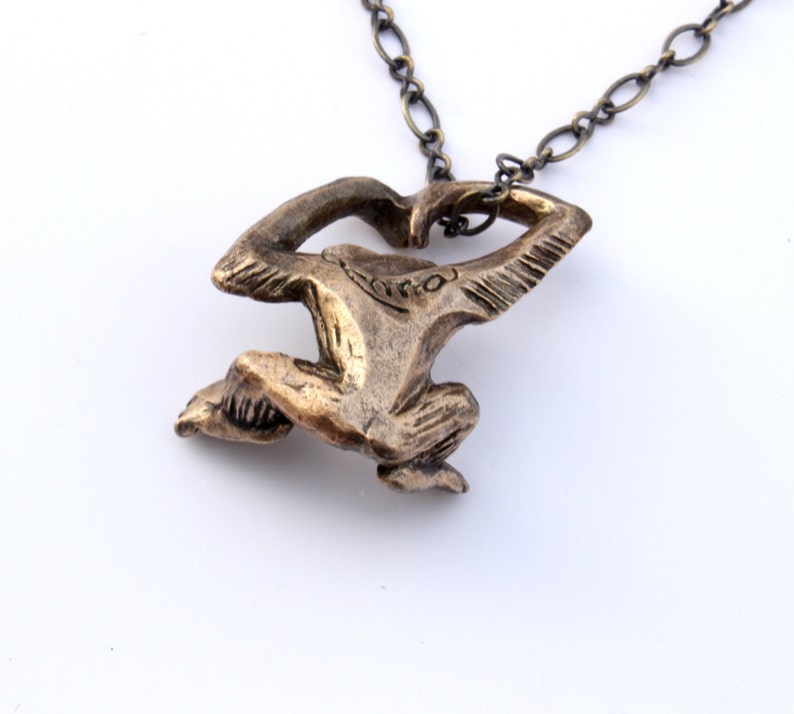 Puede incluir: Un collar con un colgante de bronce que presenta una figura estilizada de una persona en posici&oacute;n de cuclillas. El colgante est&aacute; unido a una cadena de plata.