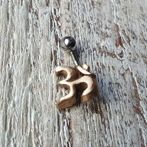 Könnte beinhalten: Ein goldener Körperschmuck-Piercing mit einem Om-Symbol und einer silbernen Kugel.