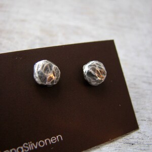 May include: A pair of silver earrings with a textured, hammered finish. The earrings are displayed on a brown card with the text "Anna Siivonen".
