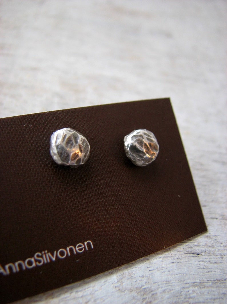 May include: A pair of silver earrings with a textured, hammered finish. The earrings are displayed on a brown card with the text "Anna Siivonen".
