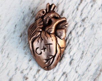 Può includere: Un ciondolo a forma di cuore color rame con un design in rilievo e le lettere "C+T" incise sul davanti.