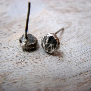 May include: A pair of silver stud earrings with textured surfaces. The earrings are on a wooden surface.