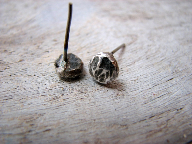 May include: A pair of silver stud earrings with textured surfaces. The earrings are on a wooden surface.