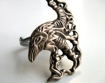 Puede incluir: Un anillo de plata con un diseño de cuervo estilizado y detallado. El cuervo está representado con las alas extendidas y la cabeza girada hacia un lado. El anillo está hecho de metal y tiene un acabado liso.