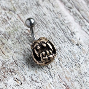 Puede incluir: Un piercing de ombligo plateado con un dije de oro detallado de un rostro de monstruo gruñendo.