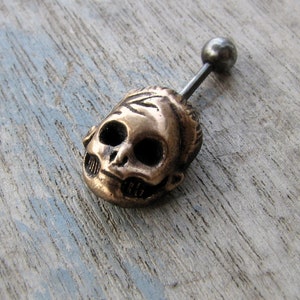 Puede incluir: Un charm de calavera dorado con una cara detallada y una barra de barbell plateada. El charm está unido a la barra con una pequeña bola plateada.