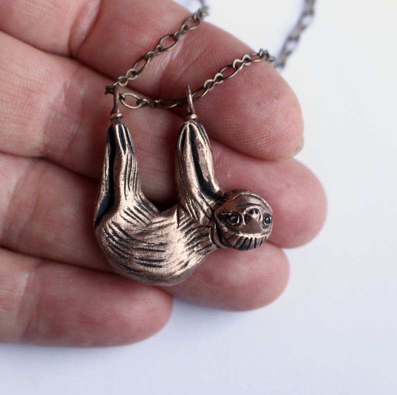 Pode incluir: Um pingente de pregui&ccedil;a de cor cobre pendurado em uma corrente prateada. A pregui&ccedil;a &eacute; representada em posi&ccedil;&atilde;o de pendurar com os bra&ccedil;os estendidos.