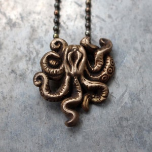 Può includere: Una collana con un ciondolo in bronzo a forma di polpo. Il polpo ha otto tentacoli ed è raffigurato in posizione arrotolata. La collana è composta da una catena con piccole perle.