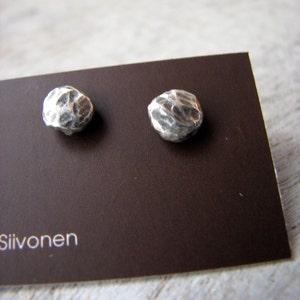 May include: A pair of silver earrings with a textured, irregular, pebble-like design. The earrings are displayed on a brown card with the text "Siivonen" printed on the bottom left corner.
