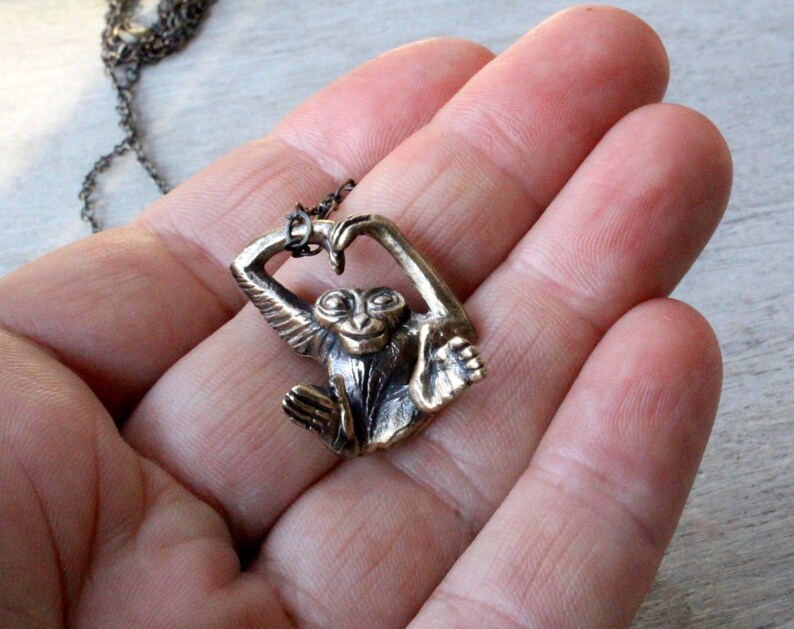 Puede incluir: Un collar de bronce con un dije de mono en forma de coraz&oacute;n. El mono est&aacute; sosteniendo sus manos juntas para formar un coraz&oacute;n.