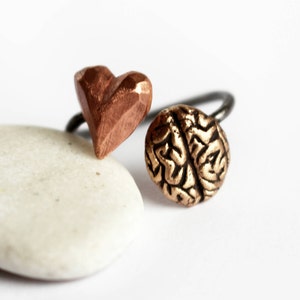 Può includere: Un anello con un cuore di rame e un ciondolo a forma di cervello dorato. L'anello è su una superficie di pietra bianca.