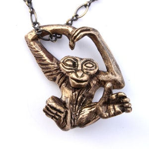 Puede incluir: Un collar con un colgante de bronce en forma de mono con los brazos levantados en forma de coraz&oacute;n. El mono tiene una cara detallada y un cuerpo texturizado.