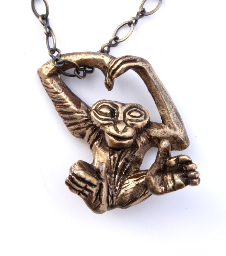 Puede incluir: Un collar con un colgante de bronce en forma de mono con los brazos levantados en forma de coraz&oacute;n. El mono tiene una cara detallada y un cuerpo texturizado.