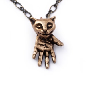 Puede incluir: Un collar dorado con un colgante con forma de gato. El gato está extendiendo su pata y tiene los ojos cerrados. La cadena es de metal.