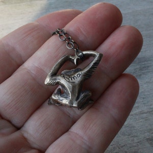 Puede incluir: Un collar de plata con un colgante con forma de persona con los brazos en alto. El colgante tiene la palabra "Anna" inscrita.