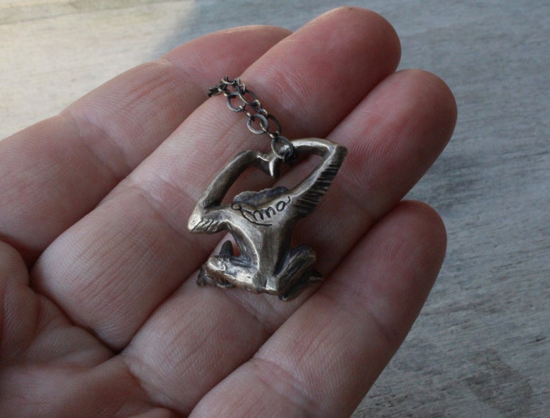 Puede incluir: Un collar de plata con un colgante con forma de persona con los brazos en alto. El colgante tiene la palabra "Anna" inscrita.