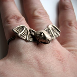 Può includere: Un anello d'argento con un design a forma di pipistrello. Il pipistrello ha le ali e un viso dettagliato.