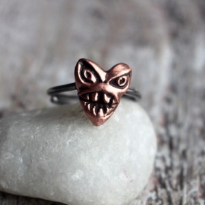 May include: A copper ring with a heart-shaped design featuring a scary face with teeth. The ring is on a white stone background.