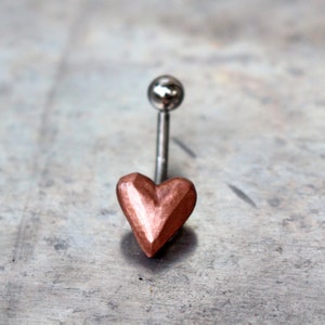 Puede incluir: Un piercing de ombligo en forma de corazón de color cobre con una barra de plata.