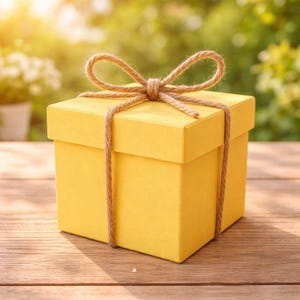 Puede incluir: Una caja de regalo amarilla brillante atada con un lazo de cuerda natural. La caja cuadrada está sobre una superficie de madera, con un fondo verde borroso. La cuerda envuelve la caja y está atada con un lazo decorativo en la parte superior.