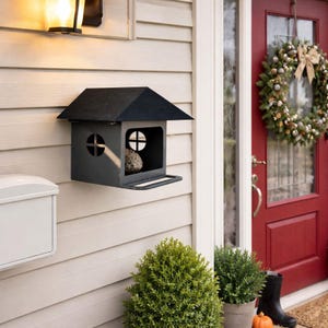 Puede incluir: Una pajarera gris oscuro con un techo negro está montada en una pared beige claro. La pajarera tiene una pequeña percha y ventanas circulares. También se ven un buzón blanco y una corona decorativa.