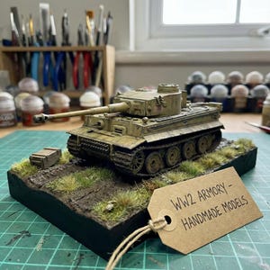 May include: A detailed model of a World War II Tiger tank, painted in olive drab and tan, sits on a diorama base with grass and dirt. The model includes a long gun barrel and intricate track details. A tag reads "WW2 Armory - Handmade Models."