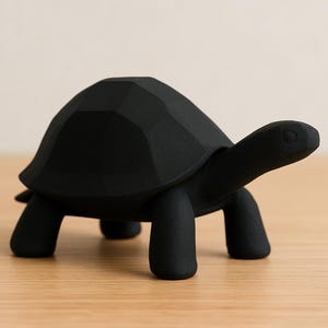 May include: A black, geometric-shaped turtle figurine. The sculpture has a faceted shell, a long neck, and four stubby legs. The turtle is set against a light-colored background and sits on a wooden surface.
