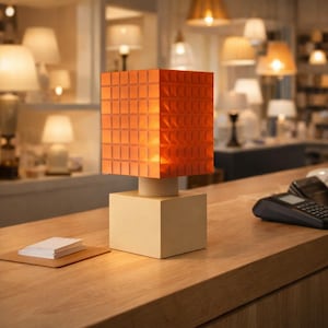 Puede incluir: Lámpara de mesa moderna con una pantalla cuadrada naranja texturizada. La lámpara se apoya sobre una base color crema y un soporte cilíndrico. El fondo muestra otras lámparas y una superficie de madera.