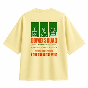 Puede incluir: Una camiseta de color amarillo pálido con un gráfico en la espalda que muestra tres iconos rectangulares verdes de un detonador, unas tijeras y un delantal. El texto dice: BOMB SQUAD, CRISIS PREVENTION UNIT, CERTIFIED SITUATION DEFUSER, EVERYONE MAKES IT WORSE, I CUT THE RIGHT WIRE.