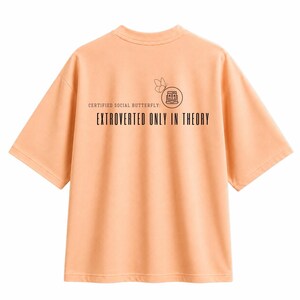 Op de afbeelding: Een perzikkleurig T-shirt met korte mouwen en een zwarte afbeelding op de achterkant. Het ontwerp bevat een vlinder, een klein computericoontje met de tekst 404 en de woorden: CERTIFIED SOCIAL BUTTERFLY: EXTROVERTED ONLY IN THEORY.