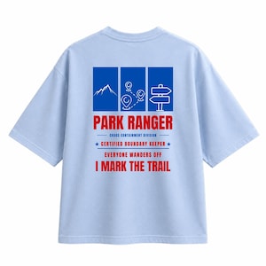 May include: A light blue t-shirt featuring a graphic on the back with three blue panels showing a mountain, location pins, and a signpost. Text reads: PARK RANGER, CHAOS CONTAINMENT DIVISION, CERTIFIED BOUNDARY KEEPER, EVERYONE WANDERS OFF, I MARK THE TRAIL.