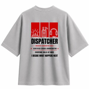 May include: A light gray short-sleeve t-shirt featuring a graphic on the back. The design includes three red squares with white icons of a siren, a radio, and a headset. Text reads: DISPATCHER, EMERGENCY COORDINATION CENTER, CERTIFIED CHAOS COORDINATOR, EVERYONE CALLS AT ONCE, I DECIDE WHAT HAPPENS NEXT.