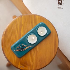 Puede incluir: Un portavelas de cerámica turquesa con dos velas encendidas, colocado sobre una mesa de madera. El soporte tiene un diseño metálico. La mesa es redonda y de madera. La imagen también muestra una silla de madera y una superficie blanca.