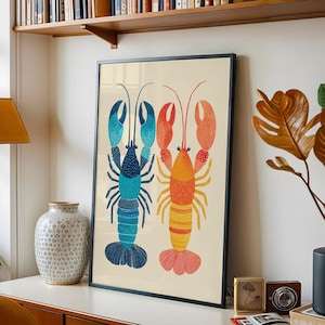 May include: A framed art print featuring two stylized lobsters. One lobster is blue and turquoise, the other is orange and yellow. The artwork has a cream-colored background and is displayed in a black frame. The print is a decorative piece.