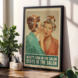 May include: Framed vintage-style print featuring two women in curlers, one whispering to the other. The print has a teal and orange color scheme with the text "WHAT'S SAID IN THE SALON STAYS IN THE SALON" in a teal box. The print is displayed on a wooden surface.