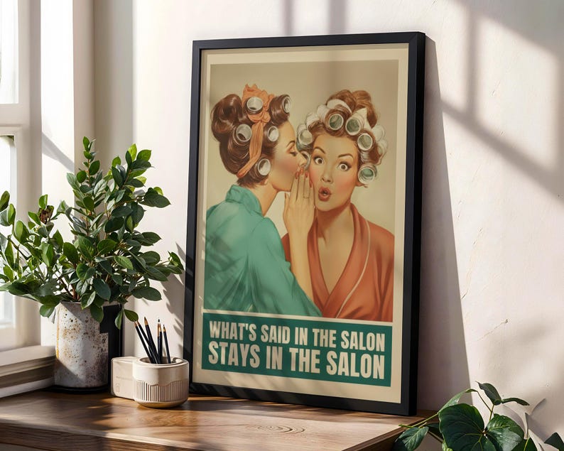 May include: Framed vintage-style print featuring two women in curlers, one whispering to the other. The print has a teal and orange color scheme with the text "WHAT'S SAID IN THE SALON STAYS IN THE SALON" in a teal box. The print is displayed on a wooden surface.