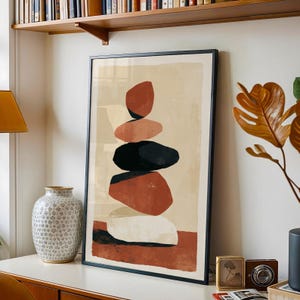 May include: A framed vertical wall art print featuring a stacked stone composition in shades of terracotta, black, and cream against a light beige background. The artwork displays five rounded, abstract shapes balanced vertically in a column.