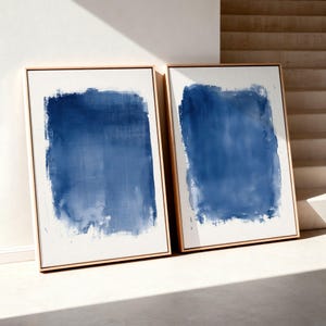 May include: Two rectangular wall art pieces in light wood frames, each featuring a central block of deep blue watercolour-style paint on a white background. The blue pigment displays varied saturation and visible brushstroke textures.
