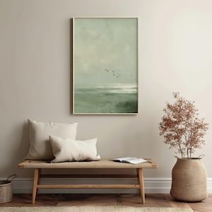 May include: A framed print depicting a tranquil seascape in muted greens and greys. A group of birds are silhouetted against the sky. The artwork is displayed above a wooden bench with cream-colored cushions. A woven basket and a decorative plant are also visible.