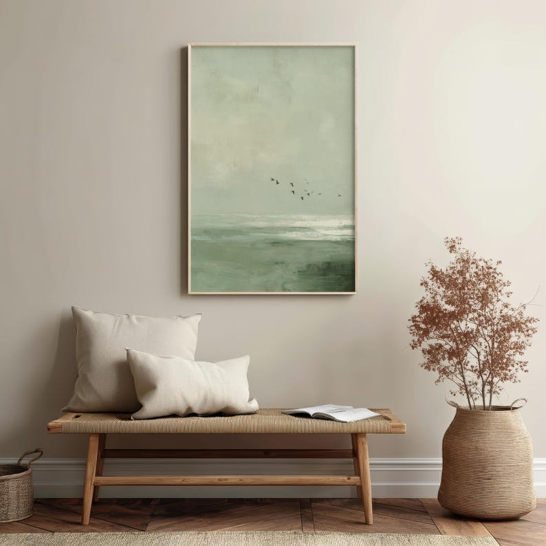 May include: A framed print depicting a tranquil seascape in muted greens and greys. A group of birds are silhouetted against the sky. The artwork is displayed above a wooden bench with cream-colored cushions. A woven basket and a decorative plant are also visible.