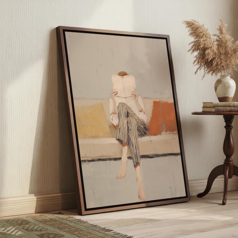 May include: A framed artwork shows a person reading a book on a sofa. The person wears striped trousers and a white shirt. The sofa has yellow and orange cushions. The artwork is in a wooden frame, leaning against a wall.