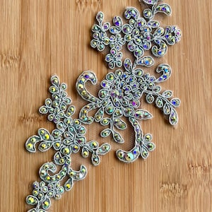 Op de afbeelding: Twee zilveren bloemenkanten applicaties met iriserende strass-steentjes. De applicaties zijn gerangschikt op een houten oppervlak. De strass-steentjes reflecteren een regenboog aan kleuren en geven het ontwerp een vleugje glans en elegantie.