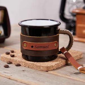 Puede incluir: Taza de esmalte negro con una envoltura y asa de cuero marrón. La envoltura de cuero presenta el nombre "Samuel" y una estrella. La taza está sobre un posavasos de corcho, con granos de café esparcidos alrededor. Una radio vintage y un molinillo de café están en el fondo.